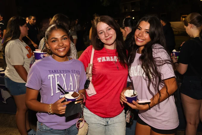 A group of people at an event, with three individuals in the foreground smiling and wearing Tarleton State University apparel. They are holding small bowls, possibly with ice cream. Other attendees and activities are visible in the background.