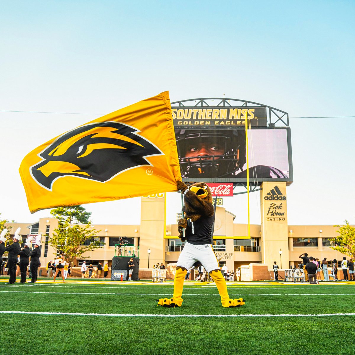 On a football field, a Golden Eagle mascot in a black jersey and white pants waves a large gold flag featuring a Golden Eagle logo. A jumbotron is in the background with people scattered around the field.