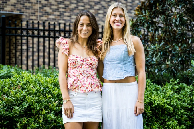 Two women smiling, standing side by side in front of a lush green hedge and a brick wall backdrop. One wears a floral top, the other a blue one, both in light skirts.