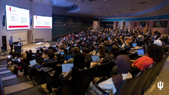 A large lecture hall filled with attendees, each with a laptop. A speaker stands at a podium near two projected screens displaying information about IU Bloomington, with text such as "Kellems Faculty and Staff" visible. The tiered seating arrangement allows everyone a clear view of the presentation.