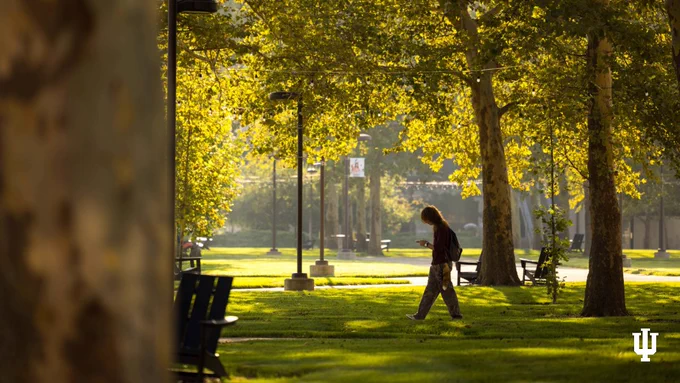 A person walks on a tree-lined path in a sunlit park. The Indiana University logo is visible in the bottom right corner.