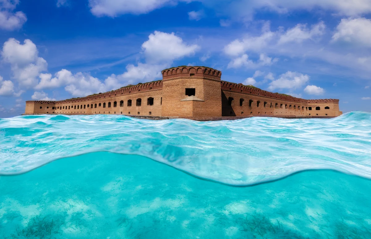 Historic brick fort surrounded by clear turquoise ocean water under a partly cloudy blue sky.