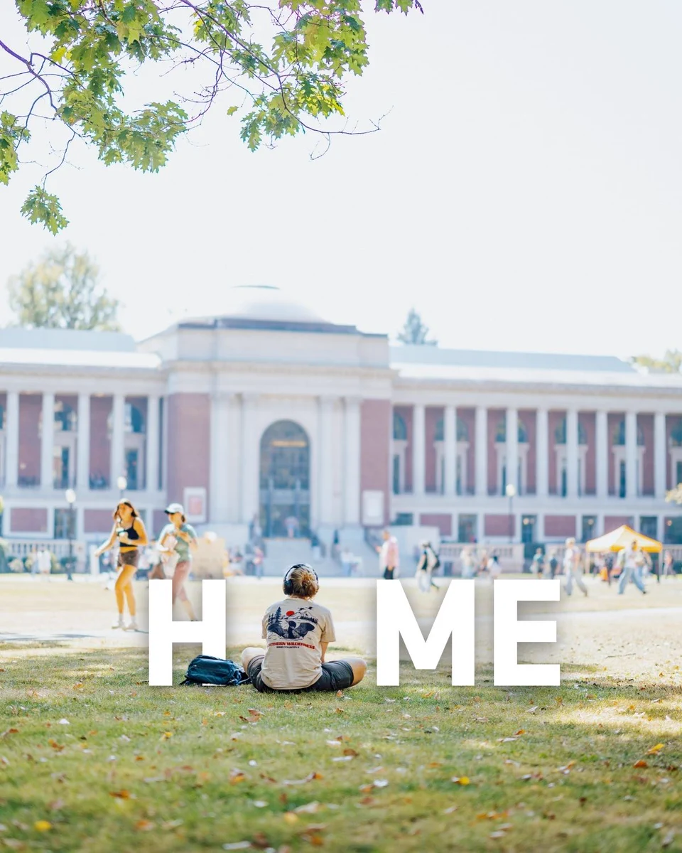 Student seated on a lawn facing the Memorial Union building while wearing headphones, with people walking across the quad and the word “HOME” overlaid on the image.