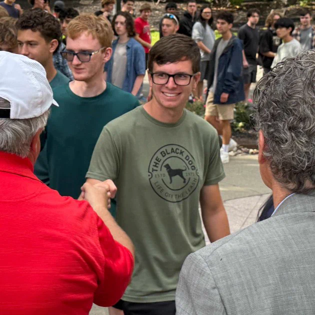 A person wearing a "The Black Dog" t-shirt smiles while shaking hands with another individual in a red shirt. There is a group of people in the background, some wearing casual attire, walking or standing around.