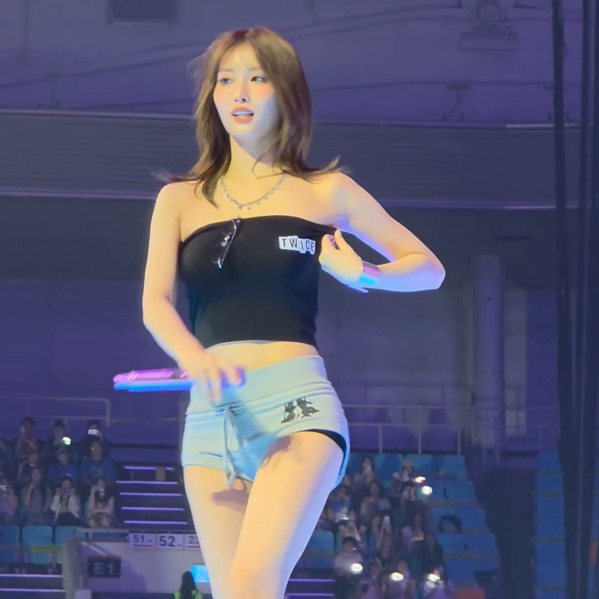 A young woman, identified as Momo from the K-pop group TWICE by the "TWICE" lettering on her black tube top, performs on stage in a light blue mini skirt, striking a dance pose as fans watch from the stadium seating below.