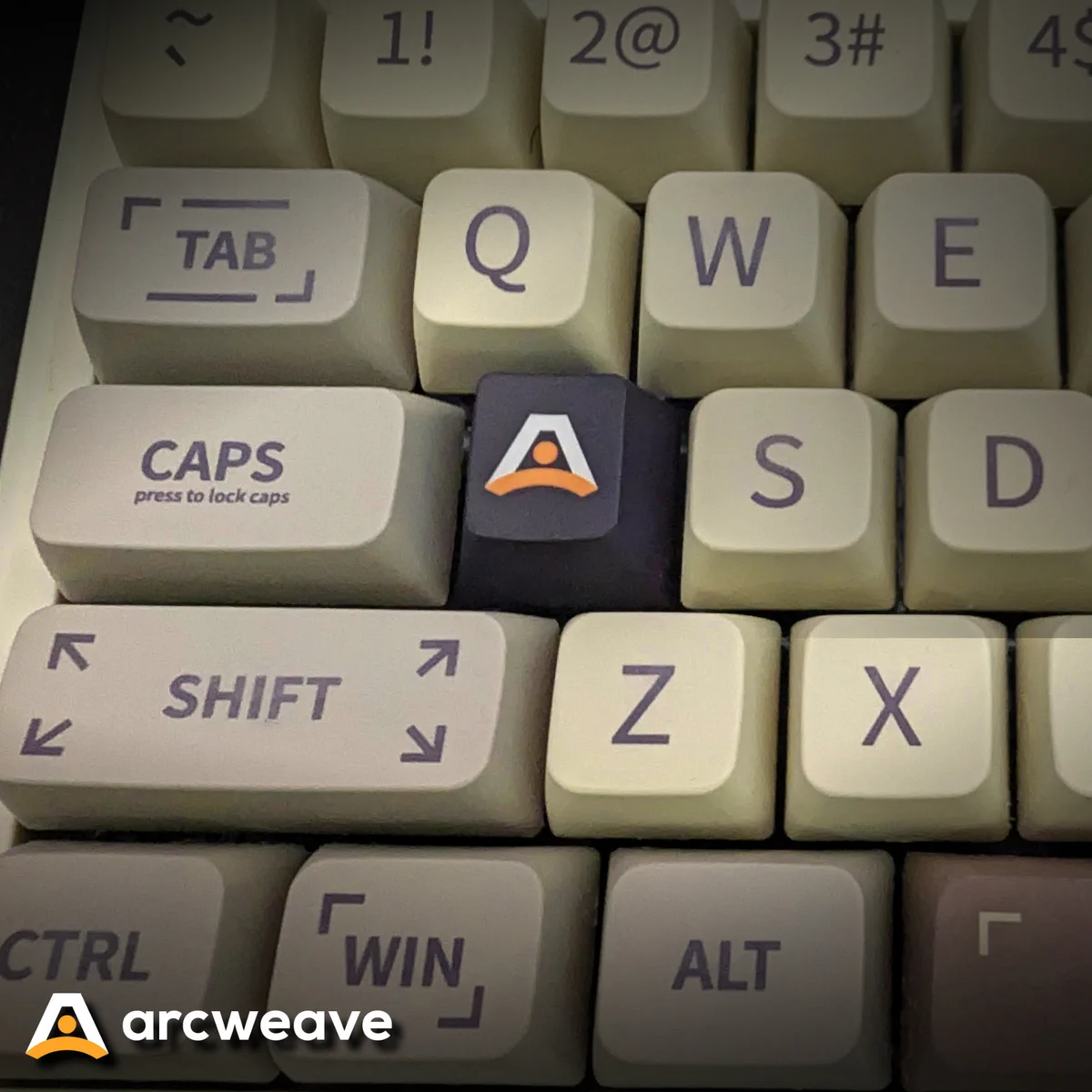 Photo of the left side of a mechanical keyboard with white keys. The A key has been replaced with a blue key that shows the Arcweave logo.
