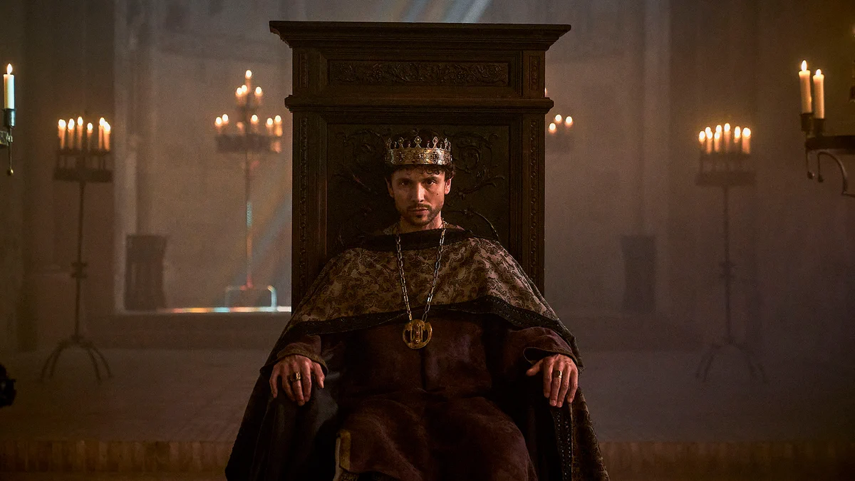 A  man in a crown sits on a wooden throne in a historical scene. The room is lit by multiple candles on large stands.