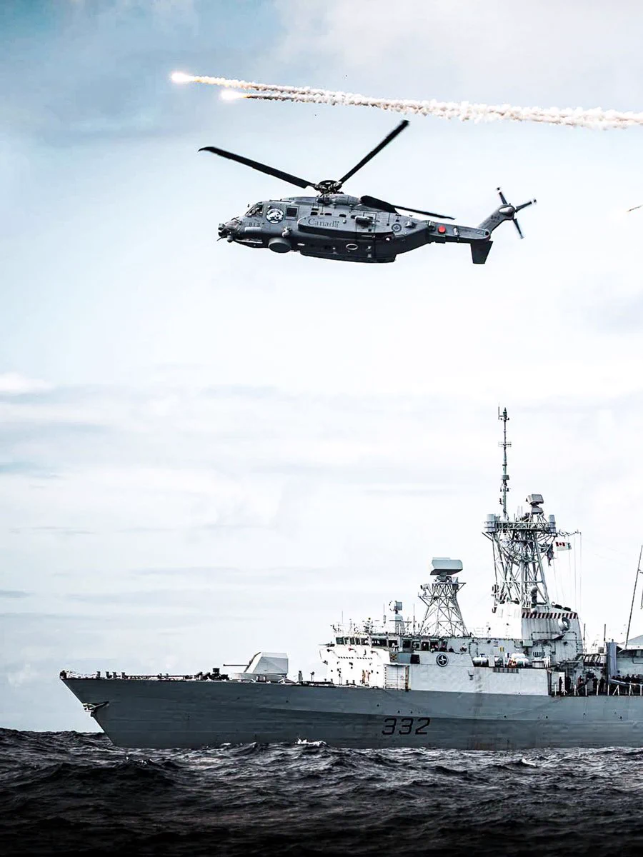 Canadian frigate HMCS Ville de Québec at sea with a helicopter hovering over