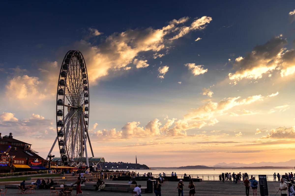 A Ferris wheel towers over a bustling waterfront pier at sunset. People gather near buildings like \"Fisherman's Restaurant\" and \"Wing Over Washington Flying Ride\". The sky is aglow with orange, pink, and blue hues, with sun rays piercing through clouds above a calm bay and distant mountains.