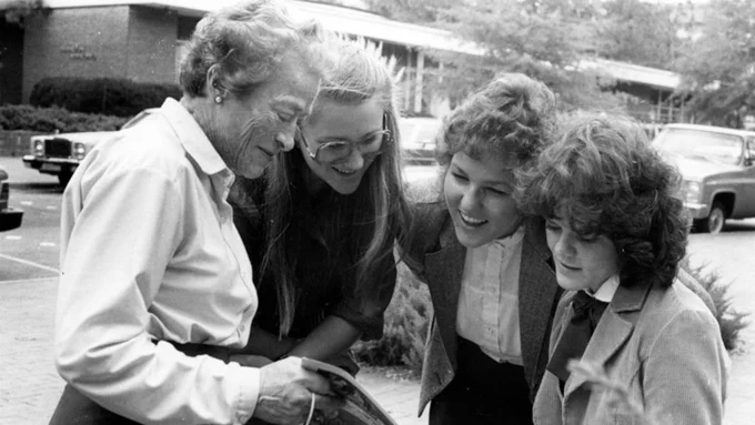 Four women of different ages closely examining a book or document outdoors in a parking lot setting.
