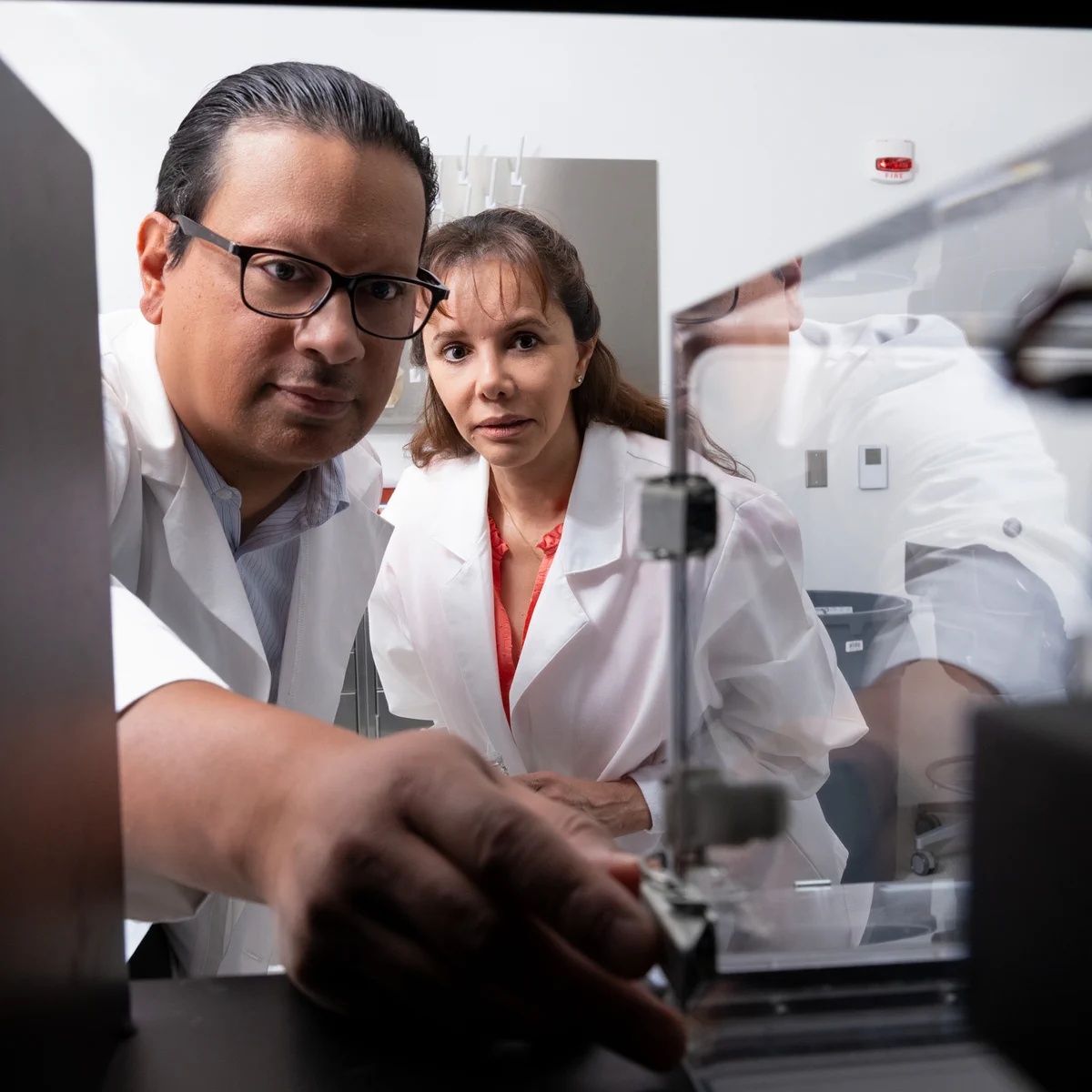 Ian Mendez, associate professor of pharmacy, and Liliana Maynez-Anchondo, a doctoral student in the Department of Psychology at The University of Texas at El Paso, wearing lab coats, work in a laboratory. Mendez adjusts equipment, and both focus intently. Various lab instruments and machines are visible in the background.