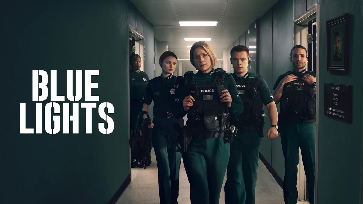 Three police officers walk down a corridor in police vests, as two more experienced officers stand in open doorways watching them. Blue Lights is written in block capital letters in white.