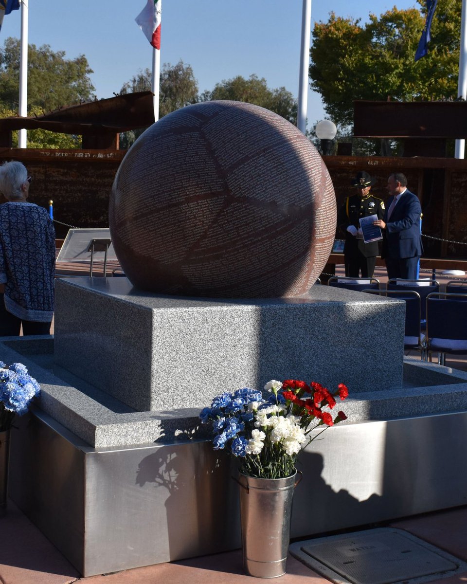 Spherical memorial engraved with names of the fallen on a stone pedestal surrounded by flowers and people at a 9/11 remembrance event.