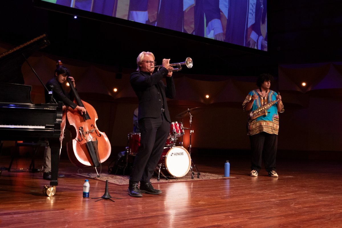 A jazz ensemble performs on a stage. A musician plays the trumpet at the front, with a double bassist, drummer, and saxophonist nearby. A piano is positioned on the left.