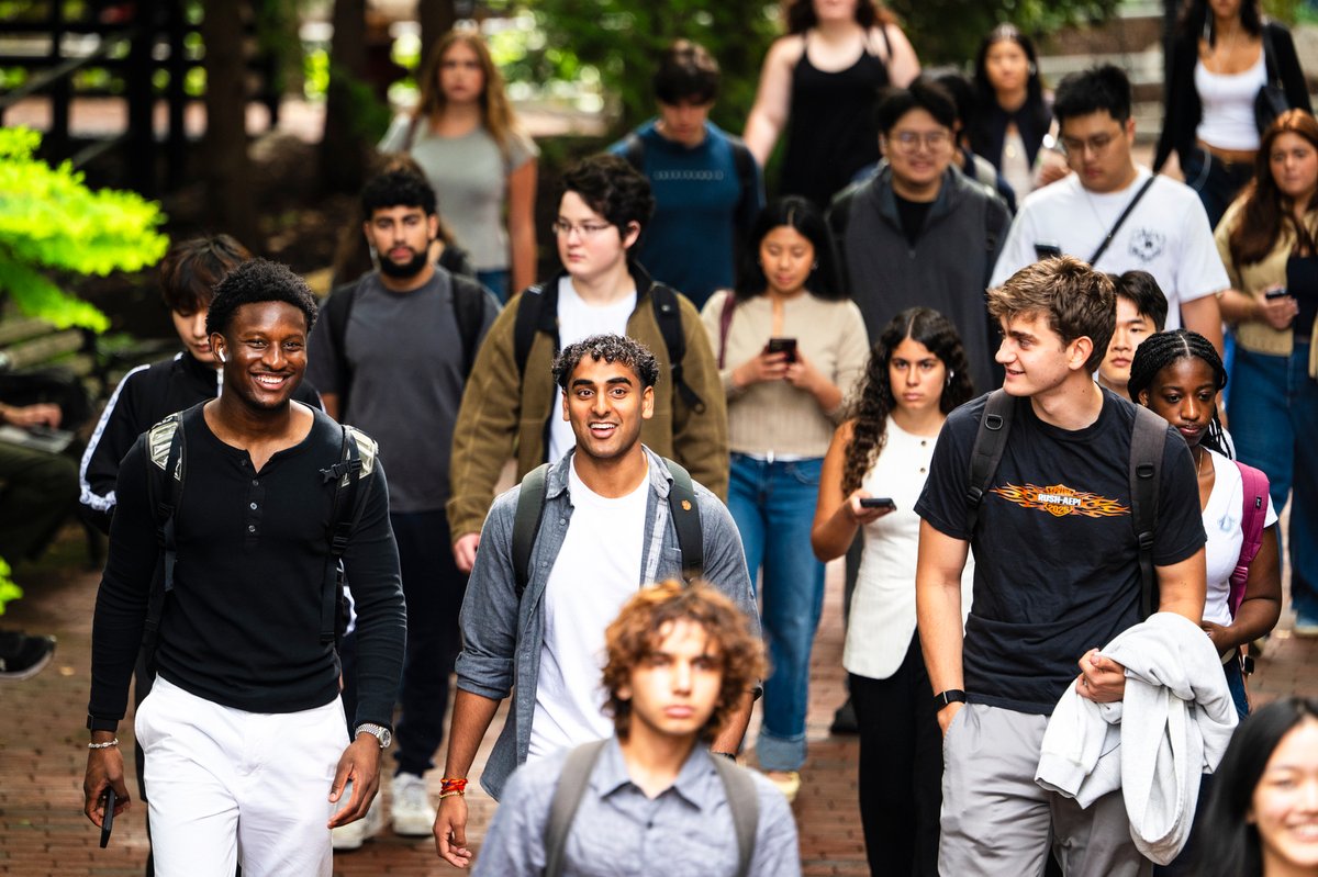 A diverse group of young adults and students walk on an outdoor pathway. Three smiling men in the foreground.