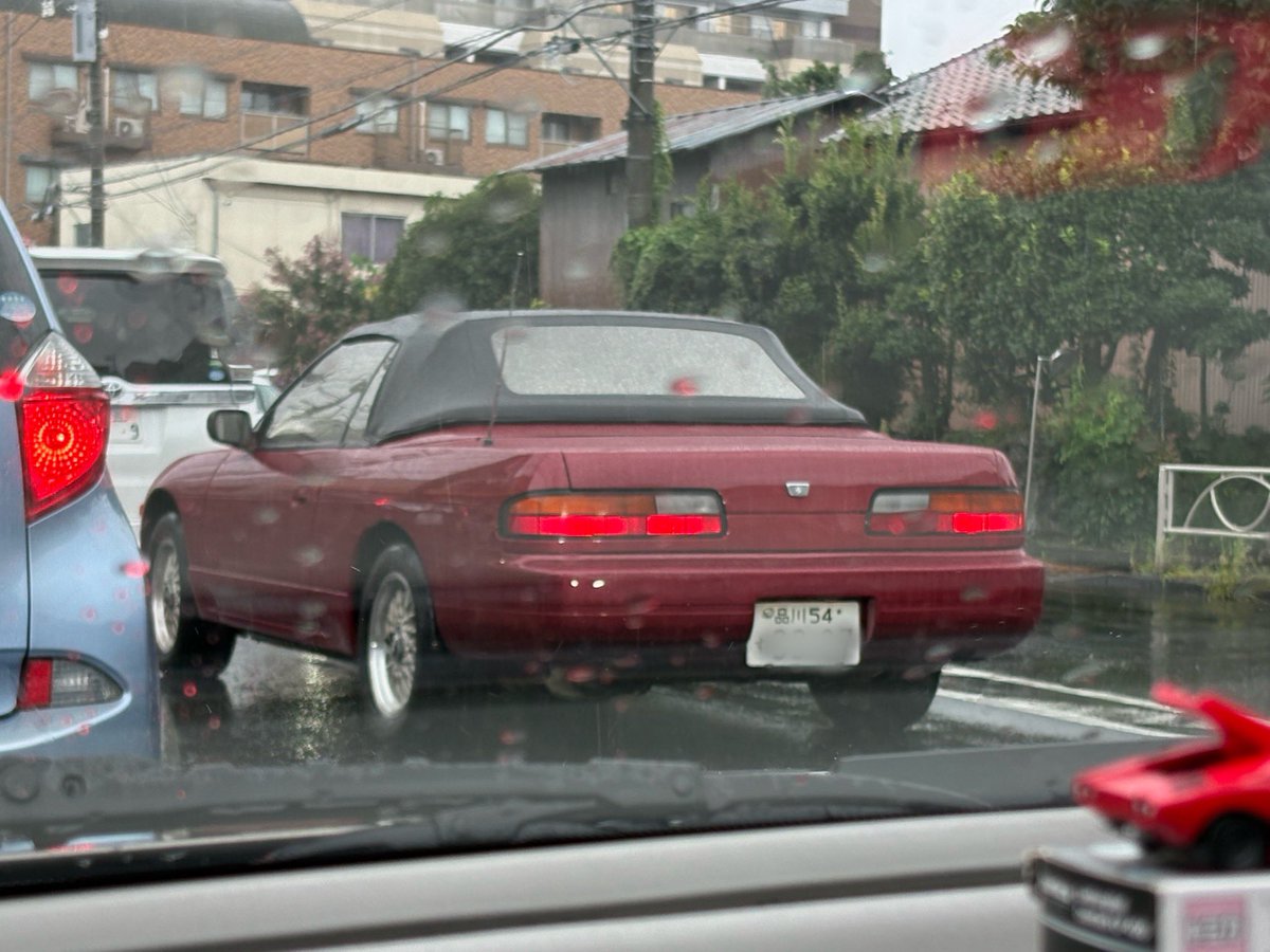 vz61_scorpion2's tweet photo. 雨の日、前を走る一台のS13シルビアに目が止まった。

まず「品川54」である。いまやS13そのものが十分に古いクルマになったが、こうして2桁分類番号を掲げたまま走っている個体となると、俄然話が違ってくる。クルマだけでなく、ナンバープレートまで当時の空気を背負っているようで、妙にありがたみがある。

ところが、よく見ると何かがおかしい。ルーフの形がいつものS13ではない。

コンバーチブルなのである。

S13といえば、低いボンネットに薄いキャビン、そして端正な3ボックスのクーペスタイルを思い浮かべる。ところが屋根を幌にしても、このクルマは意外なほど様になっている。むしろ長いボンネットと低いウエストラインが強調されて、普通のシルビアとはまた違った洒落っ気が出ている。

そして、このボディカラーがまたいい。赤というより少しくすんだワインレッド、あるいはローズ系とでも言いたくなる色で、黒い幌との組み合わせが実に上品だ。若者向けスペシャリティカーだったS13が、急に大人びたGTカーのように見えてくる。

雨粒の付いた「品川54」、黒いソフトトップ、そしてこの落ち着いた赤。派手に改造しているわけでもないのに、そこらの最新スポーツカーよりよほど目を引く。

S13シルビアは売れたクルマだから、昔はいくらでも見かけた。しかし、30年以上の時を経て、2桁品川ナンバーを付けたコンバーチブルが雨の東京を普通に走っているとなれば、これはもう別格である。

こういうクルマに偶然出会うと、「古いクルマはやっぱりいいねえ」と言いたくなる。
