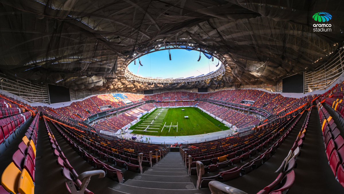 لقطة بانورامية واسعة من المدرجات العلوية تستعرض التصميم الداخلي الحديث لاستاد أرامكو (aramco stadium)، وتبرز مقاعد الجماهير المتدرجة بألوان العنابي والبرتقالي والأصفر المحيطة بالملعب بالكامل، مع سقف الاستاد المبتكر بفتحته البيضاوية المطلة على السماء الزرقاء وتدلي حوامل الإضاءة، وظهور شاشات العرض العملاقة في الأركان وأرضية المستطيل الأخضر المزودة بوحدات إضاءة وصيانة العشب، مع شعار الاستاد في الزاوية العلوية.