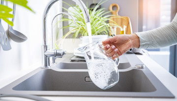 A hand is filling a clear glass pitcher with water from a modern kitchen faucet. Nearby are kitchen utensils, a potted plant, and a wooden cutting board on the countertop.