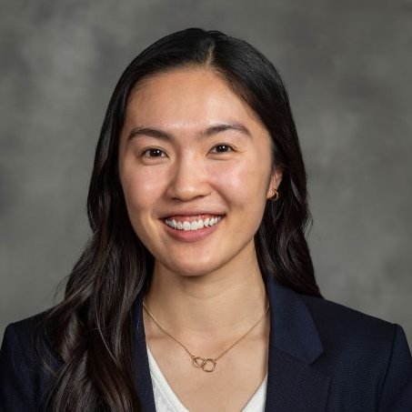 Professional headshot of Dr. Joanna Lum smiling at the camera, wearing a navy blazer over a white top and a delicate necklace, against a gray background.