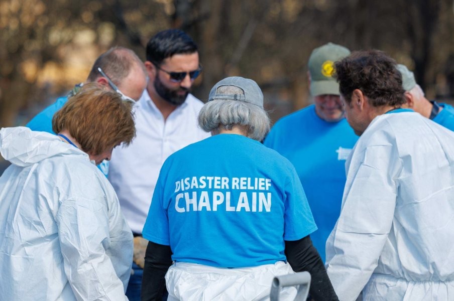 People wearing blue disaster relief chaplain shirts and white protective gear assembled in a focused group outdoors.