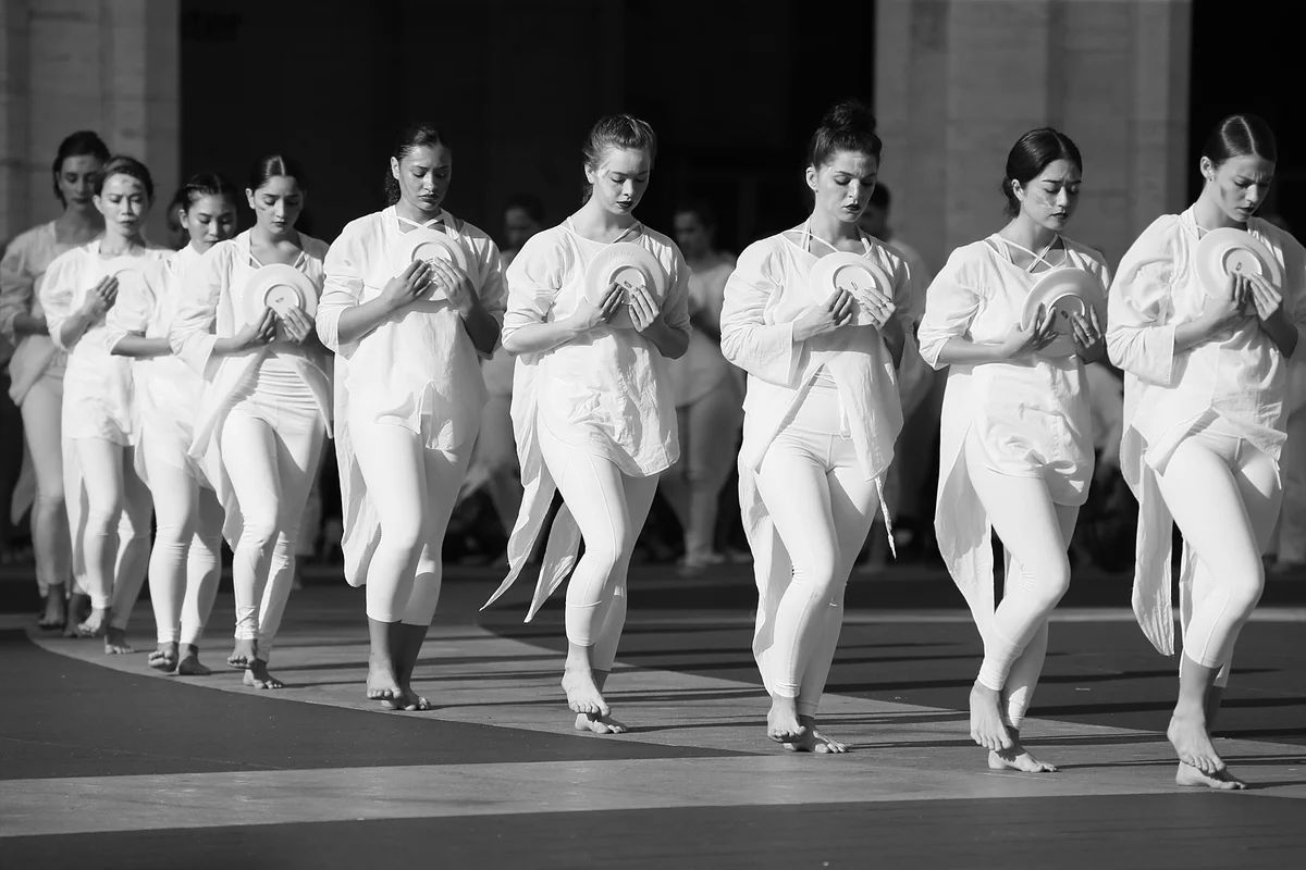 A group of individuals dressed in white, performing a coordinated dance routine on Josie Robertson Plaza. They are in a line and moving in unison, each holding a round plate.