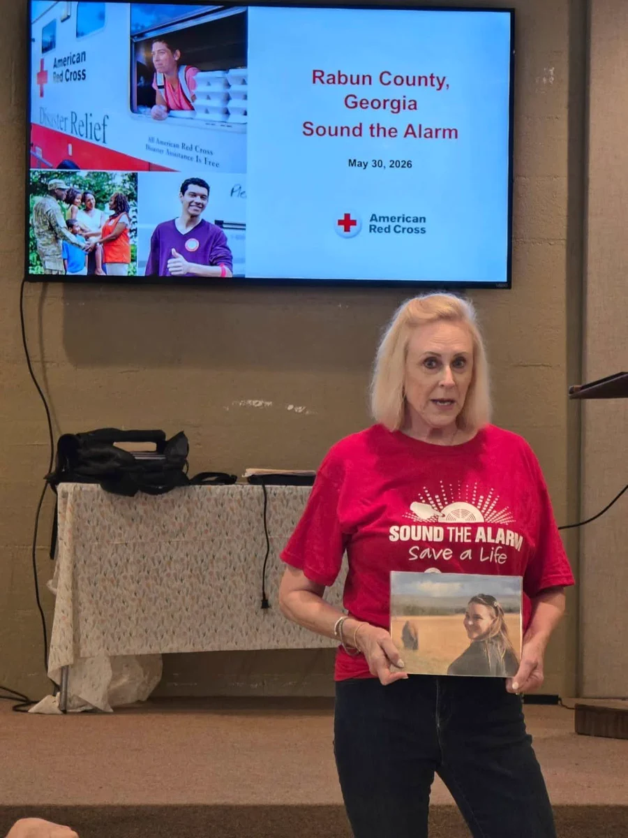 Person in a red "Sound The Alarm" T-shirt is holding a photo of her daughter and standing in front of a screen displaying "Rabun County, Georgia Sound the Alarm, May 30, 2026" with the American Red Cross logo.