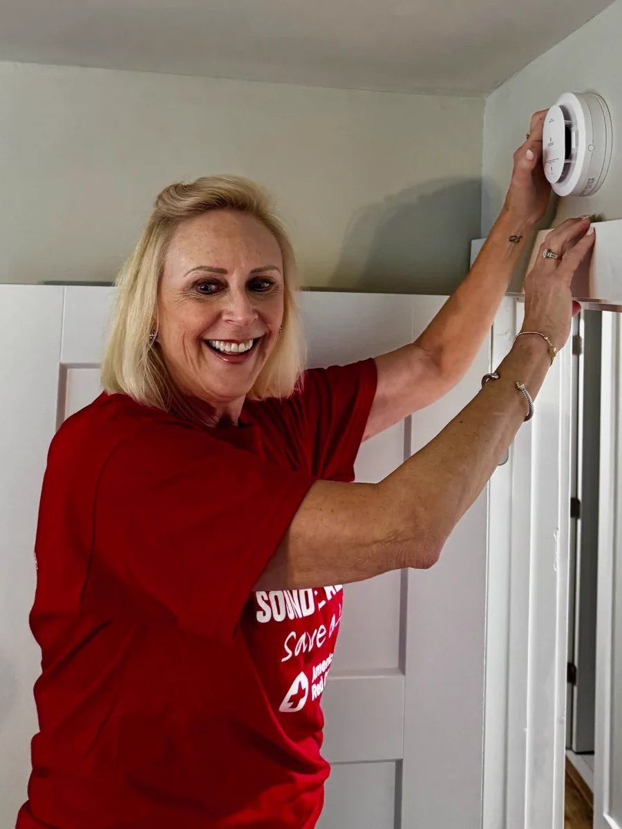 A person wearing a red American Red Cross shirt is installing a smoke detector on a wall inside a room. They are smiling and reaching up to secure the device.