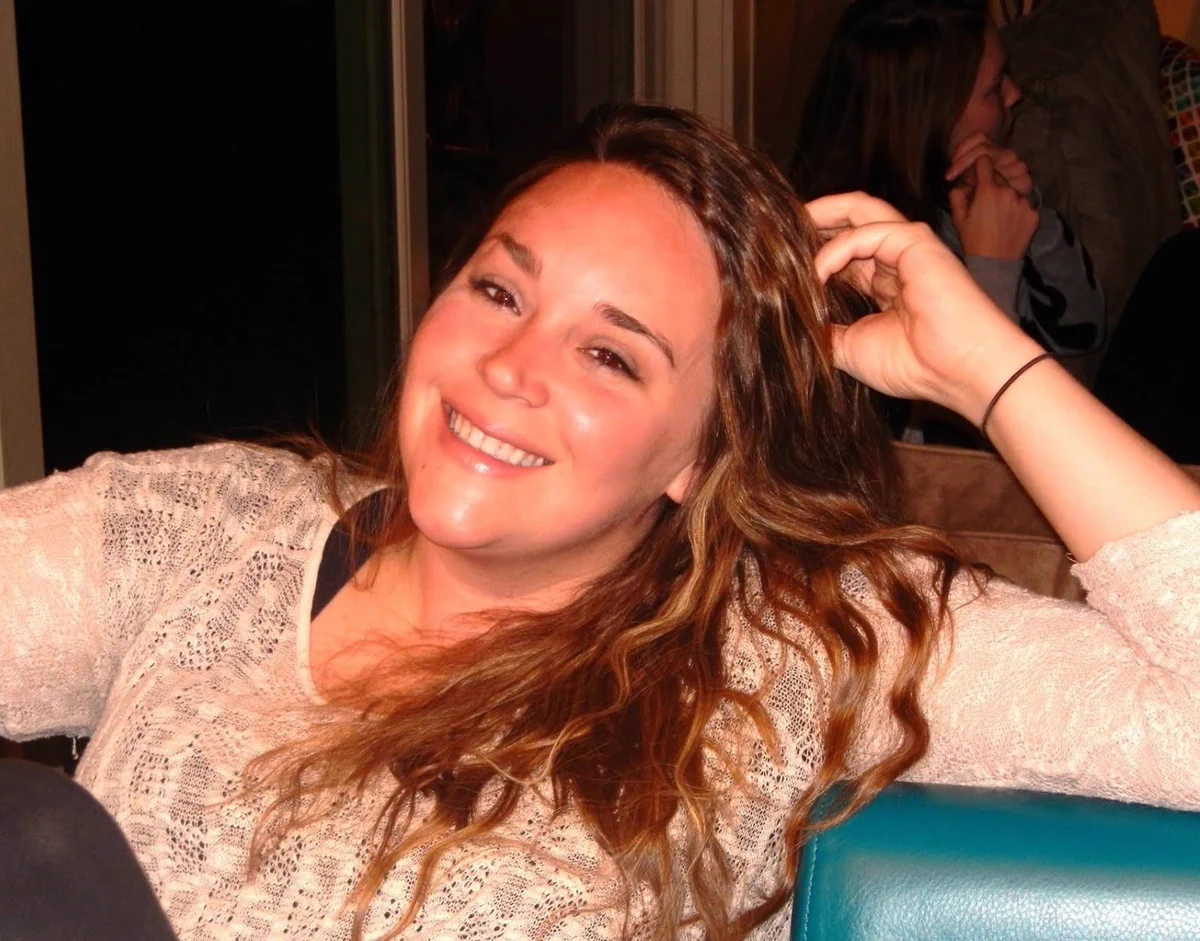 A woman posed for a picture smiling with her hand  clasping her head. She has long brown hair and a long white shirt. 