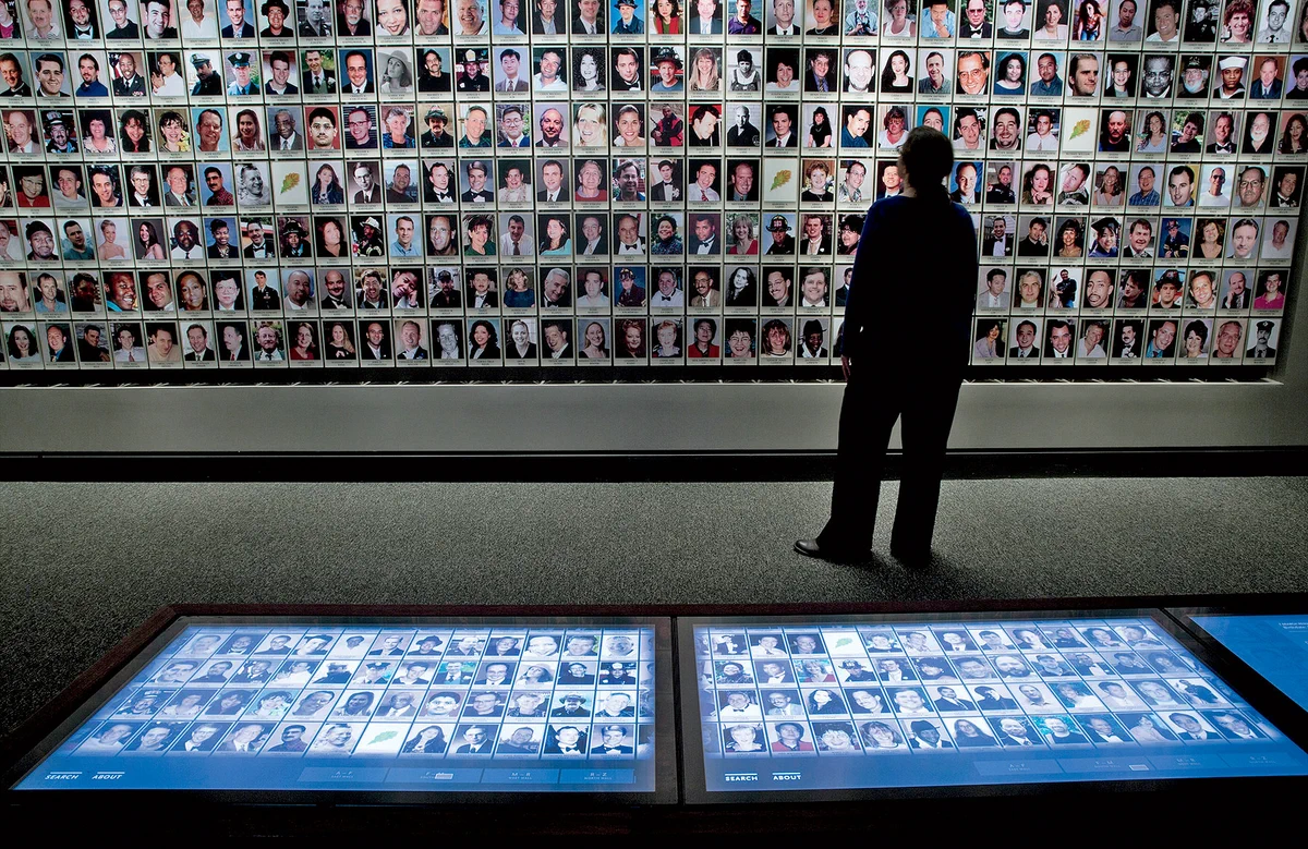 A person stands in front of a large wall displaying numerous photographs of individuals at The National 9/11 Pentagon Memorial. Two touch-screen displays are visible in the foreground, showing more images.
