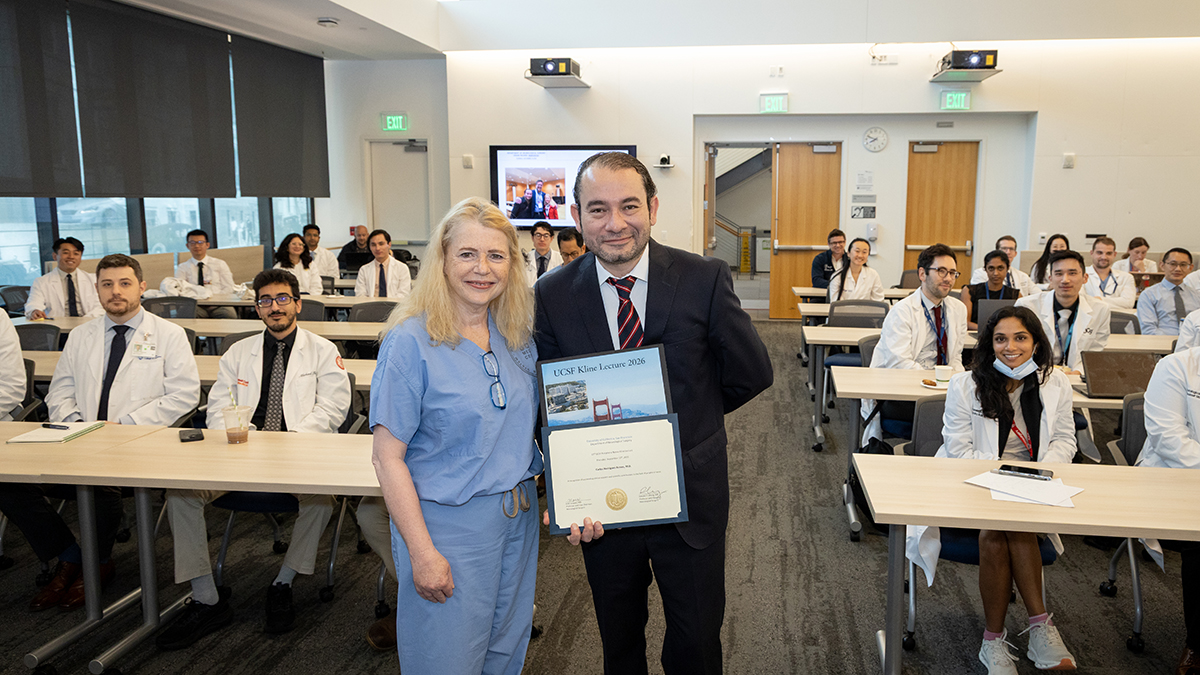 UCSF neurosurgeon Line Jacques, MD (left), stands next to guest speaker Carlos Rodriguez Aceves, MD, who is holding up an award certificate for presenting the 10th UCSF Peripheral Nerve Kline Lecture. The medical students, residents, and faculty who attended the lecture are seated behind them in the classroom.