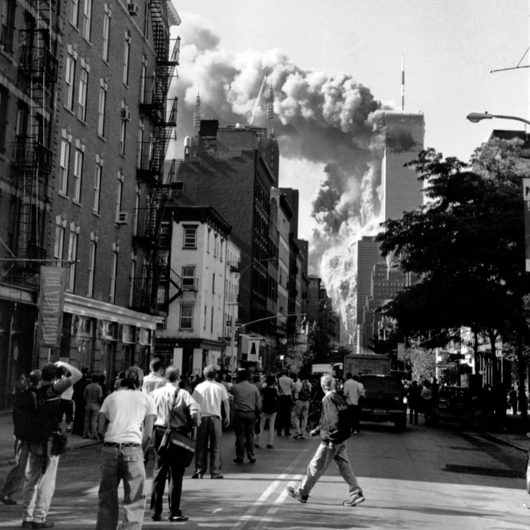 Black-and-white photograph of pedestrians in Manhattan watching smoke billow from the World Trade Center towers, conveying shock and confusion.