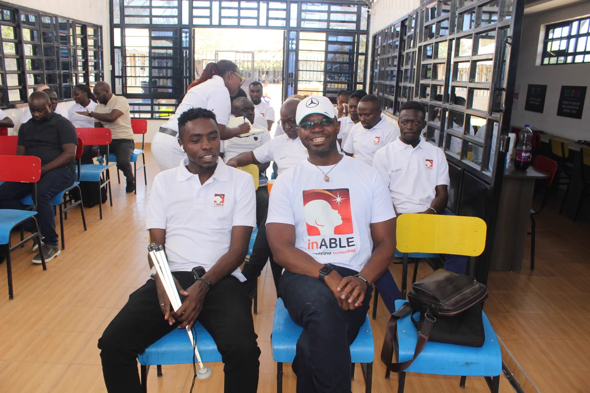 Interior view of a training session at the inABLE Digital Hub. In the foreground, Julius sits on a blue chair wearing a white inABLE polo shirt and holding a folded white cane, while Charles sits beside him wearing a white inABLE T-shirt and white cap featuring the Mercedes-Benz logo. Around them, participants wearing inABLE-branded shirts are seated on colorful chairs in a bright, open learning space.