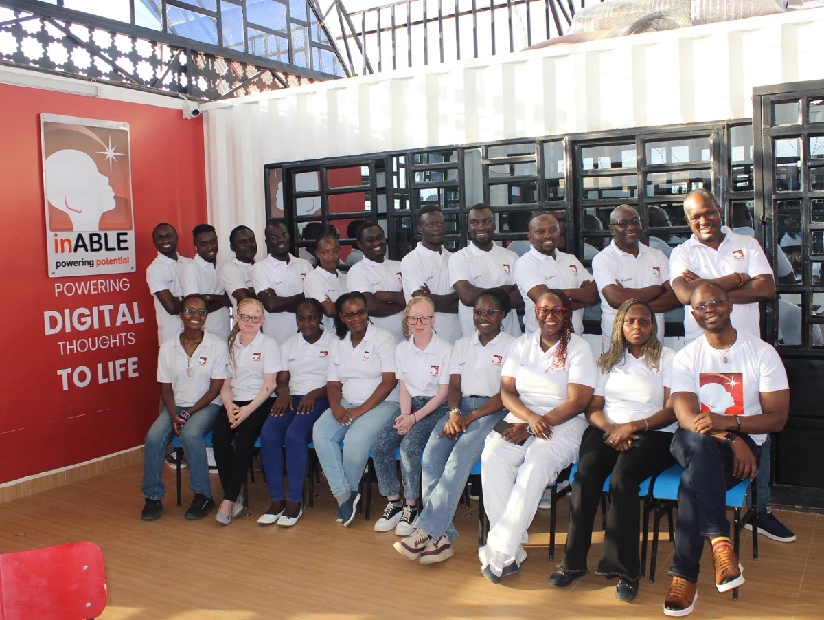 Group photo of inABLE staff gathered inside the inABLE Digital Hub. Team members, including Charles, Diana, Davis, Julius, Linda, Eva, George, Anthony, Valarie, and other staff, are arranged in two rows wearing white inABLE-branded shirts. The group is seated and standing beside a large red wall featuring the inABLE logo and the words “Powering Digital Thoughts to Life.” Bright natural light fills the modern training space through large glass-panel windows and doors.