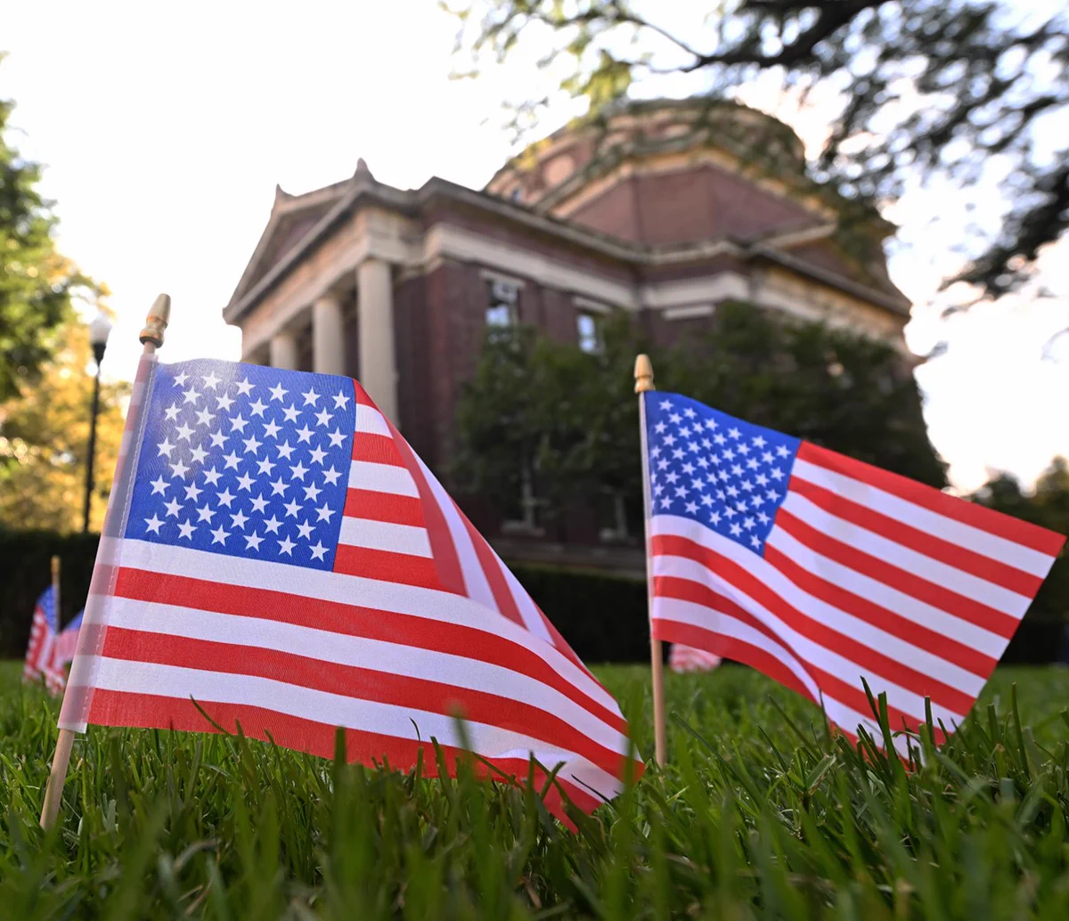 Small American flags are planted in the grass with a historic brick building in the background, featuring columns and a domed roof at Columbia University in the City of New York. The scene is lit by soft, natural light.