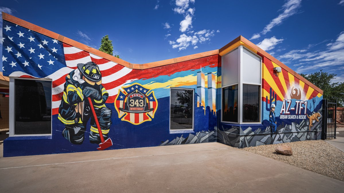 The image shows a vibrant mural on a building facade depicting a firefighter in gear, an American flag, and the Arizona Task Force 1 (AZ-TF1) emblem. The design incorporates elements of the Arizona state flag and commemorates the bravery of first responders.