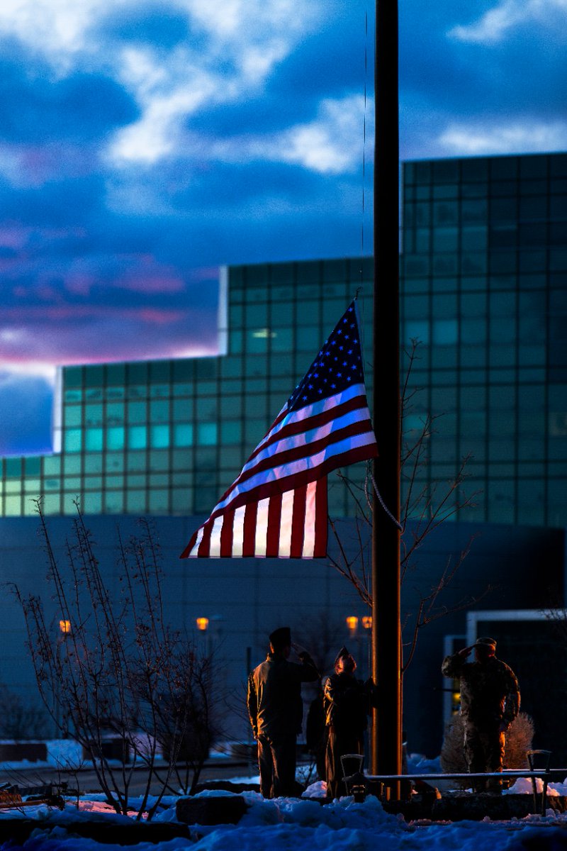 Three military personnel salute a half-mast American flag at sunrise with a modern glass building in the background.