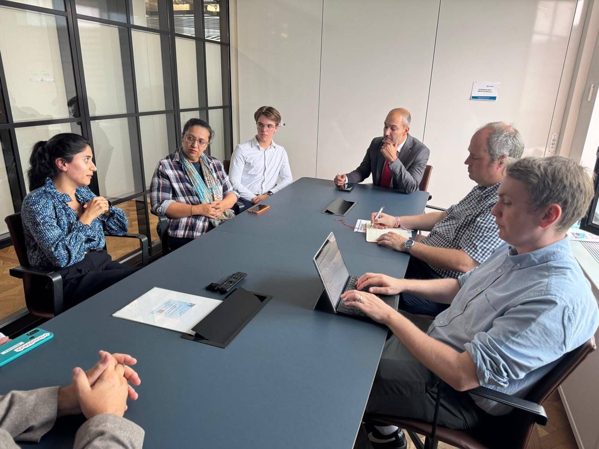 Six colleagues sit around a conference table in a modern glass-walled meeting room, one woman speaking while others listen attentively—one person types on a laptop and another takes notes.