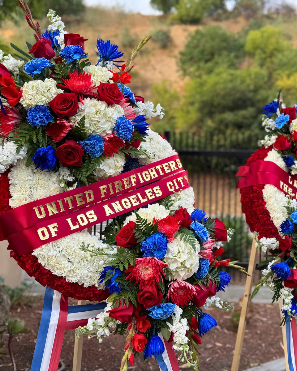 Red, white, and blue floral wreath with a ribbon reading “United Firefighters of Los Angeles City,” displayed outdoors.