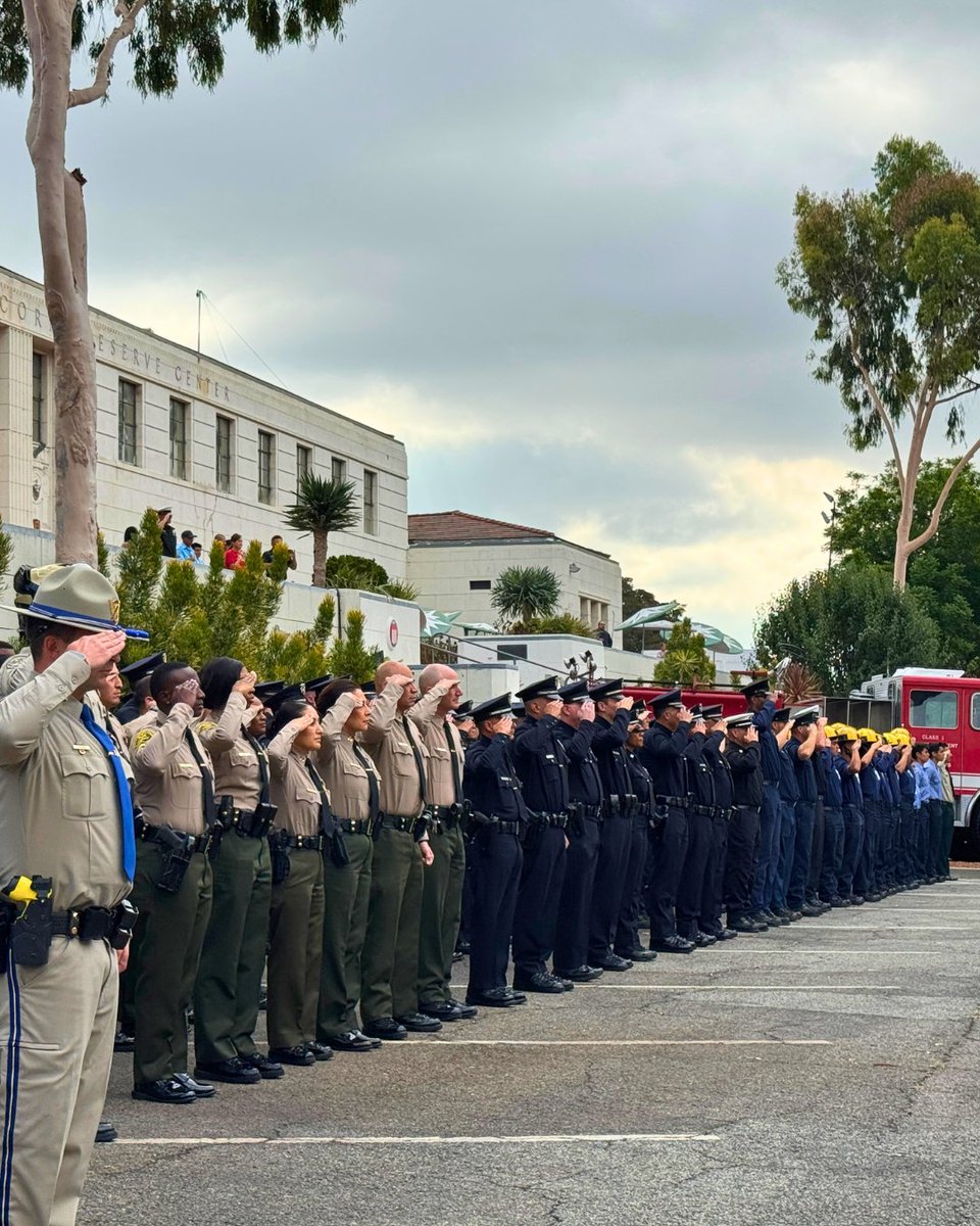 Uniformed first responders stand in a long formation, saluting together outside a civic building beside a red fire engine.