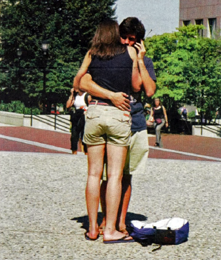 Two people are hugging outdoors on a paved area with greenery and buildings in the background. A bag is placed on the ground nearby.