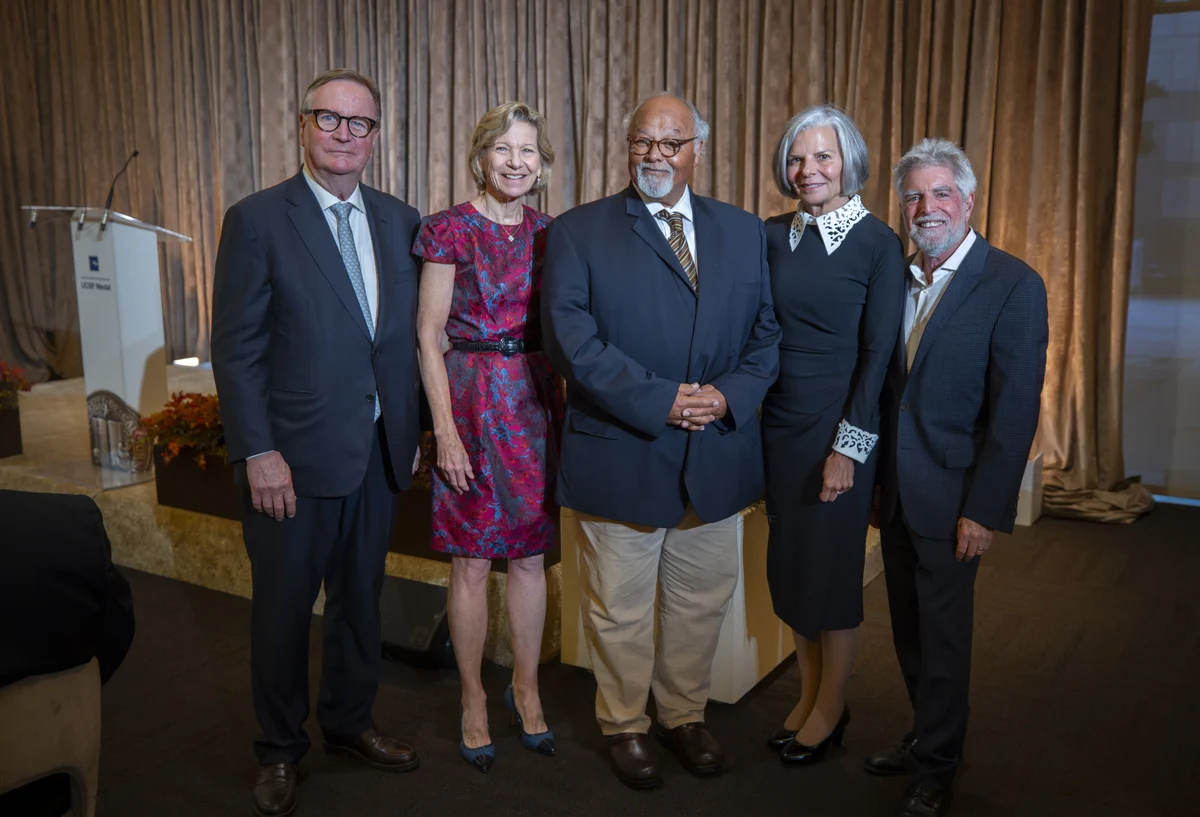 Five professionally dressed individuals standing together in front of a beige curtain at a formal event venue.