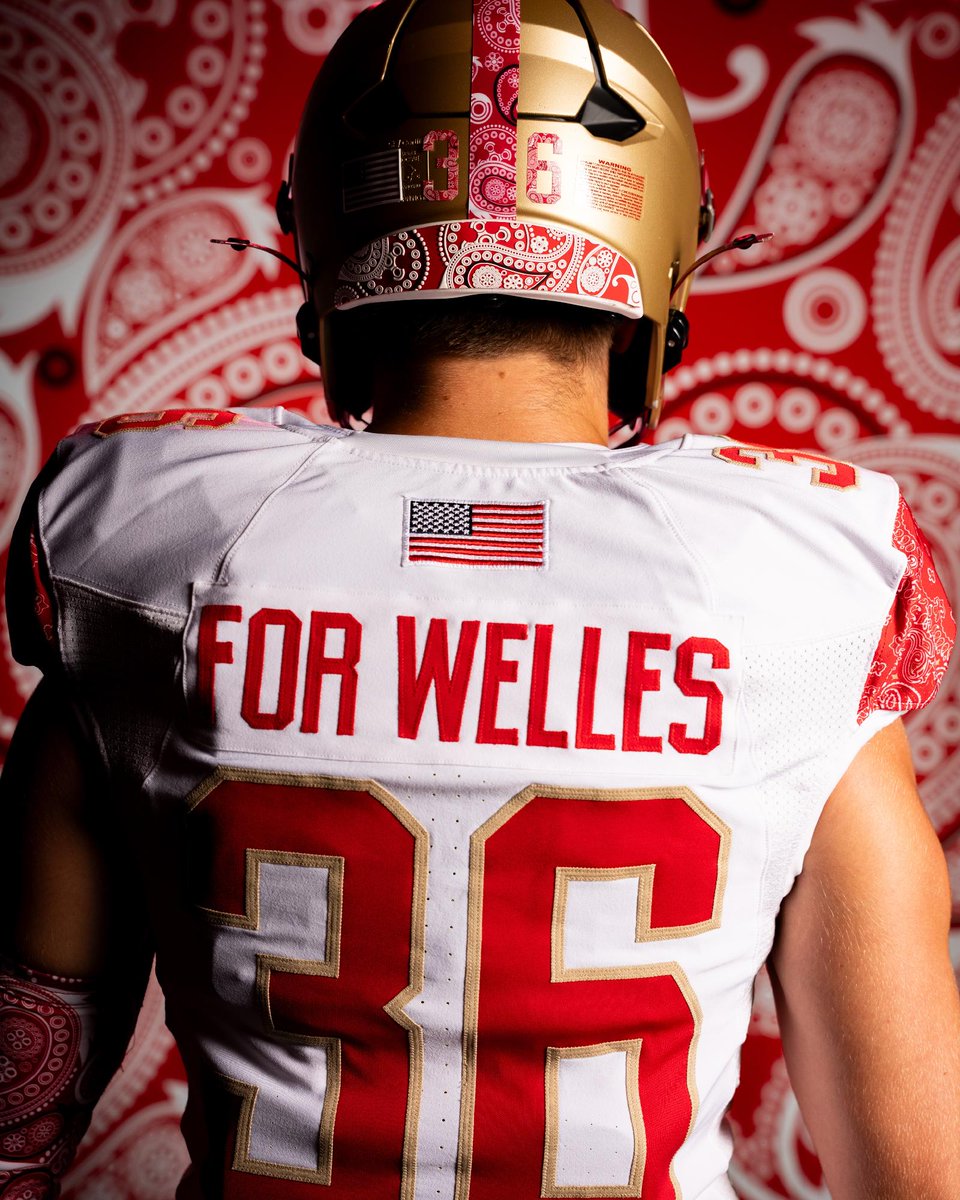 A football player is facing away, wearing a jersey with "FOR WELLES" and number 36. The helmet and uniform feature red and gold colors with paisley patterns. An American flag patch is on the jersey. The background is a red and white paisley design.