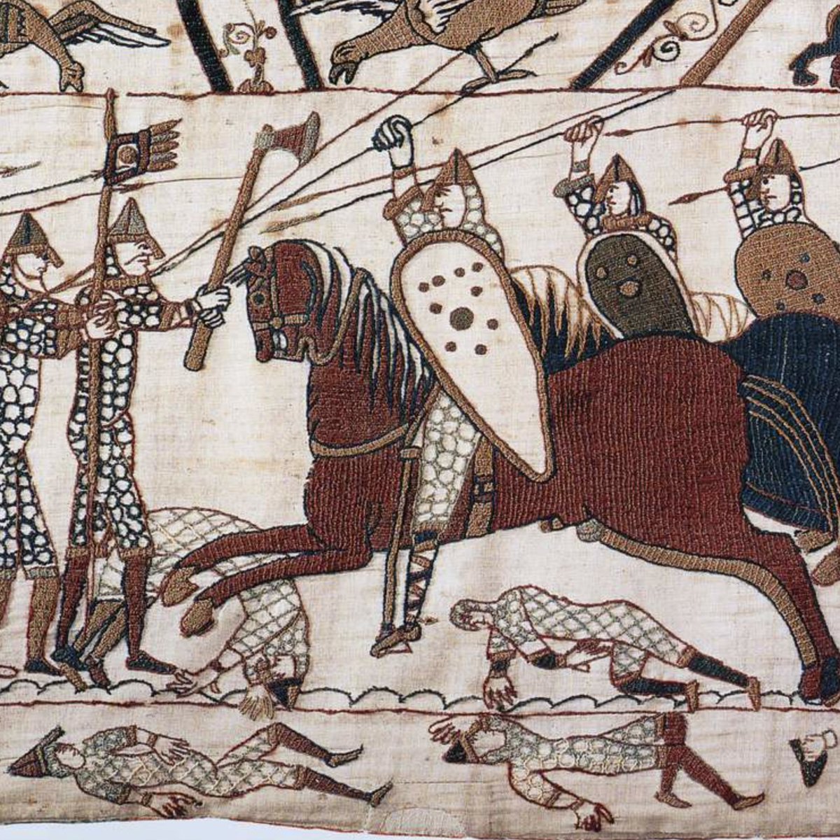 A dramatic battle scene from the Bayeux Tapestry featuring cavalrymen in chainmail riding galloping horses with raised spears and shields on the right, and on the left, two soldiers - one holding an axe and the other a flagpole. Fallen soldiers in armour lie on the ground beneath the horse's hooves.