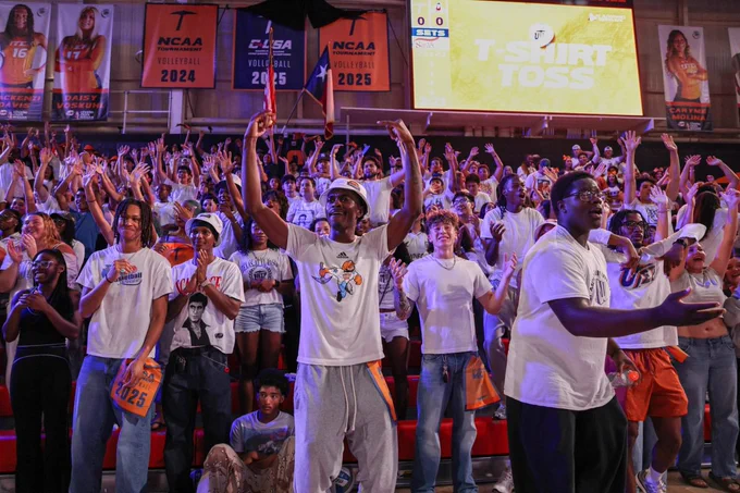 Crowd of enthusiastic fans in UTEP's Memorial Gym, many wearing white shirts. Banners for the NCAA and Conference USA Volleyball hang overhead. A large screen displays "T-SHIRT TOSS.
