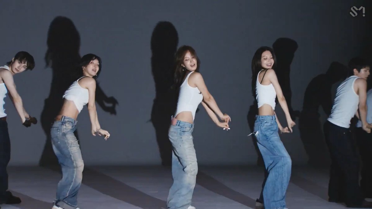Five dancers wearing white tank tops and jeans perform a choreographed routine in a studio with a dark backdrop, casting long shadows behind them.