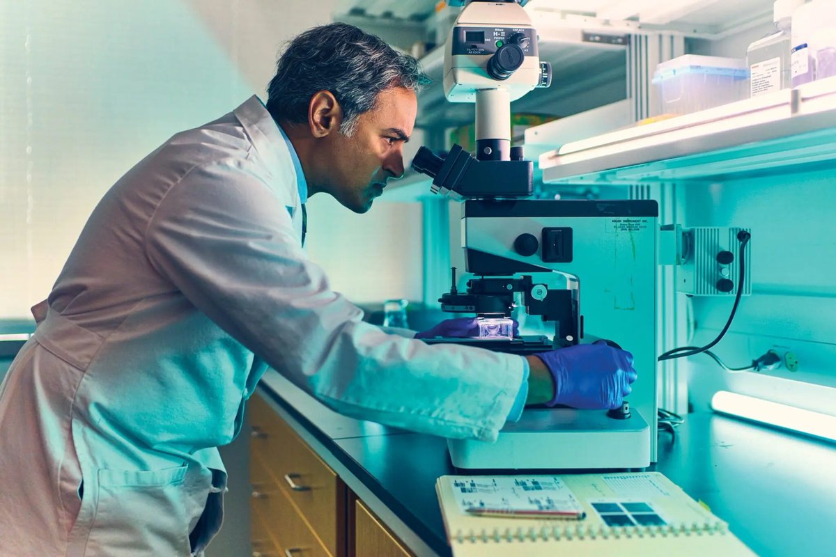 Researcher in a white lab coat and blue gloves leans in to look through a microscope at a lab bench. A camera is mounted on the microscope, an open notebook with printed data sits in the foreground, and the scene is lit in cool teal tones.