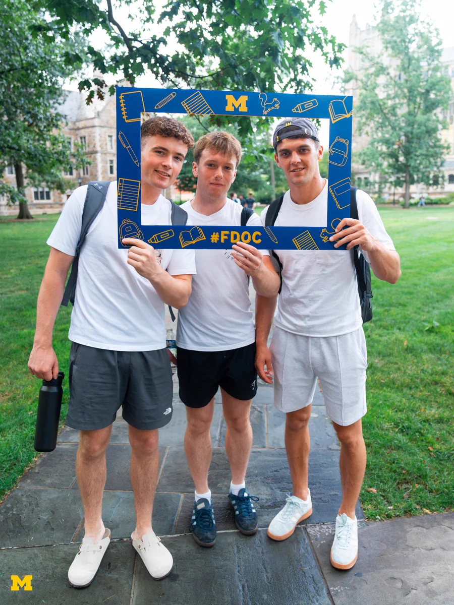 Three students on the Law Quad taking a photo with a Michigan-themed first day of classes frame.