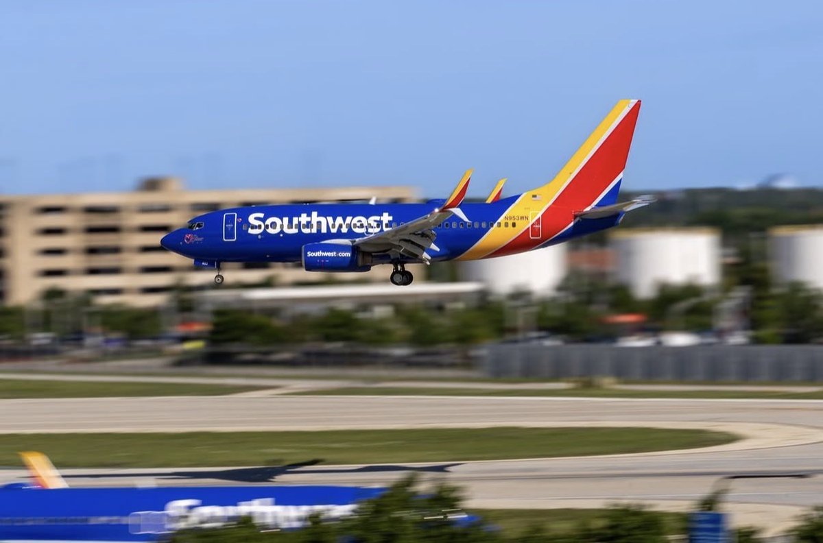 A Southwest Airlines jet is in motion, flying low over an airport runway. The plane is painted in the airline's blue, yellow, and red colors. In the background, there are blurred buildings and a clear blue sky.