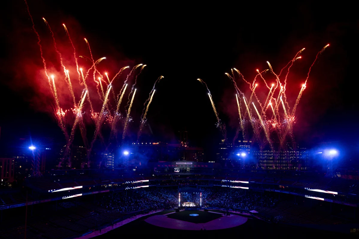 Wide shot taken of the fireworks during the Bare Knuckle Fighting Championship finale at Fenway Park. A boxing ring is seen on the middle of the field and the in-stadium lights are shining blue. 