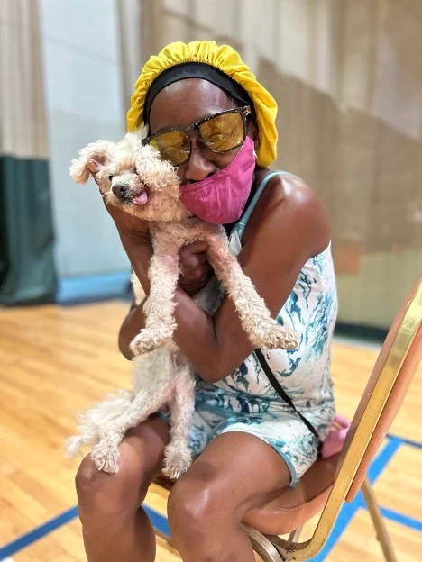 A person sitting on a chair lovingly hugs a small, curly-haired dog. They are wearing a yellow head covering, large sunglasses, and a pink face mask. The setting appears to be indoors, with a wooden floor.